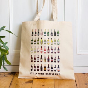 May include: A beige canvas tote bag featuring a design of various wine bottles with faces. The bottles are arranged in rows and come in different colours, including red, white, and pink. The text "IT'S A WINE-DERFUL LIFE" is printed at the bottom of the bag.
