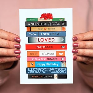 May include: A greeting card featuring a stack of book spines with titles like "AND STILL A Total" and "Happy Birthday". The spines are various colors, and a red bow adorns the top. The card is held up.