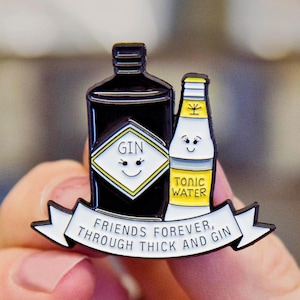 May include: A black and yellow enamel pin featuring a cartoon gin bottle and tonic water bottle with smiling faces. The pin has a white banner with the text "Friends Forever, Through Thick and Gin".
