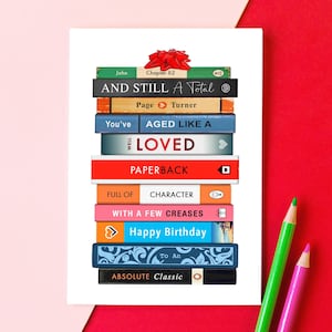 May include: A birthday greeting card featuring a stack of books with various titles and a red bow. The titles include "AND STILL A Total", "AGED LIKE A", and "Happy Birthday". Green and pink pencils are in the background.