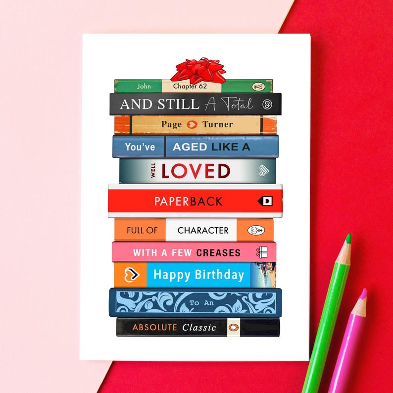 May include: A birthday greeting card featuring a stack of books with various titles and a red bow. The titles include "AND STILL A Total", "AGED LIKE A", and "Happy Birthday". Green and pink pencils are in the background.