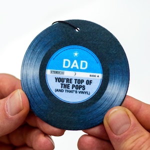 May include: A circular gift tag designed to look like a vinyl record. It is dark blue with a light blue label that reads "DAD" and "YOU'RE TOP OF THE POPS (AND THAT'S VINYL)". It has a black string attached.