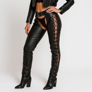 May include: Black faux leather chaps featuring a high-waisted design with a cutout front section, silver-toned metal eyelets, and black lace-up detailing running down the outer sides of each leg, paired with a matching black leather jacket.