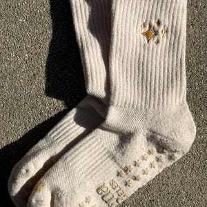 May include: A pair of off-white ribbed socks with a gold star design embroidered near the cuff. The soles feature small, star-shaped grips for traction. The text "Tina Belles" is printed on the sole of each sock.