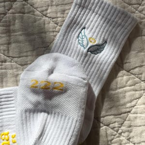 May include: White ribbed socks with embroidered angel wings and a halo in shades of blue and yellow. The number "222" is printed in yellow on the toe. The socks are made of a soft, ribbed material.