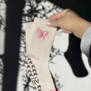 May include: A pair of cream-colored socks with a ribbed texture. Each sock features a pink embroidered butterfly near the top and a pattern of small black butterflies along the sole. The text "Ali Los Angeles" is printed on the sole.