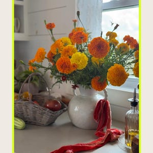 K&ouml;nnte beinhalten: Eine wei&szlig;e Keramikvase mit leuchtend orangefarbenen und gelben Ringelblumen. Ein rotes Band liegt auf der Theke. Ein Weidenkorb mit Produkten und eine Glasflasche mit &Ouml;l sind ebenfalls zu sehen.