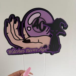 Può includere: Un cake topper viola e nero con la scritta "Witches Never Age". Il design include una mano che tiene una sfera di cristallo con il numero 40 e un gatto nero con un cappello da strega. Due bastoncini bianchi sono attaccati in basso.