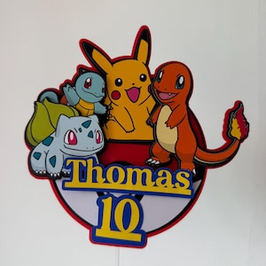 Puede incluir: Adorno de pastel colorido con los personajes Pikachu, Charmander, Squirtle y Bulbasaur. El adorno muestra el nombre "Thomas" y el número "10" en amarillo y azul. Los personajes están sobre un fondo rojo y blanco.