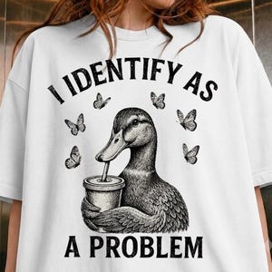 May include: A white short-sleeved t-shirt featuring a black ink-style illustration of a duck holding a cup with a straw. The text "I IDENTIFY AS A PROBLEM" is printed in black above and below the duck, surrounded by small butterfly graphics.