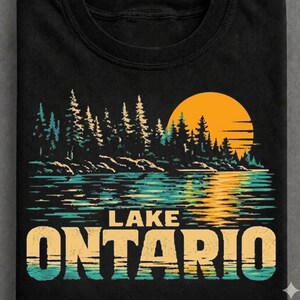 Peut inclure: Un t-shirt noir avec une impression graphique représentant une silhouette de forêt devant un grand soleil orange, avec un lac reflétant la lumière. Le texte LAKE ONTARIO est imprimé en grandes lettres grasses et claires en bas du motif.