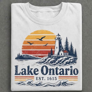 Peut inclure: Un t-shirt blanc avec une illustration représentant un phare sur une côte rocheuse avec des pins, un grand soleil couchant orange et jaune, et des oiseaux. Le texte Lake Ontario EST. 1615 est inscrit au-dessus de vagues bleues stylisées.