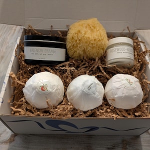 Puede incluir: Una caja de regalo llena de productos para el baño y el cuerpo. La caja contiene una vela negra con la etiqueta "Valencia Orange", una esponja de mar natural, un tarro con la etiqueta "Rosewood-Bliss" y tres bombas de baño envueltas. La caja está llena de papel marrón.