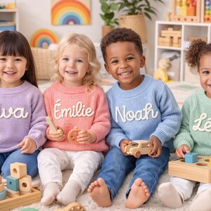 Puede incluir: Cuatro niños sonriendo con suéteres personalizados en morado, rosa, azul y verde. Cada suéter tiene un nombre bordado en blanco. Están sentados en una alfombra con juguetes de madera.
