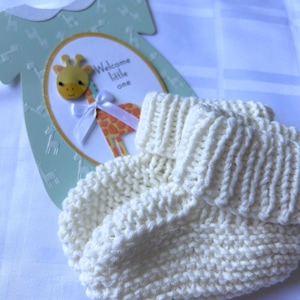 May include: A pair of cream-coloured hand-knitted baby booties with a ribbed cuff, positioned in front of a light green card shaped like a onesie featuring a giraffe illustration and the printed text Welcome little one.