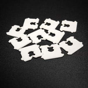 Puede incluir: Un conjunto de clips blancos de plástico para bolsas de pan, dispersos sobre una superficie negra. Cada clip tiene forma rectangular con una abertura distintiva en forma de ojo de cerradura en el centro, para sellar bolsas de pan.