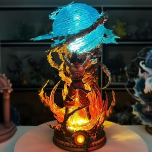 May include: A detailed figurine of a muscular character in a dynamic pose, surrounded by fiery orange and yellow flames. Above, a glowing blue energy sphere hovers. The figure is wearing orange pants and a blue sash. The base has a circular design.