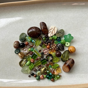 May include: A collection of assorted beads in various shades of green, brown, and amber. The assortment includes smooth round beads, faceted glass pieces, carved wooden shapes, leaf-shaped charms, and small floral-patterned components in mixed sizes.