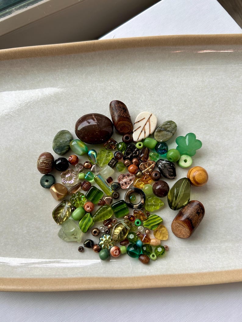 May include: A collection of assorted beads in various shades of green, brown, and amber. The assortment includes smooth round beads, faceted glass pieces, carved wooden shapes, leaf-shaped charms, and small floral-patterned components in mixed sizes.