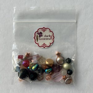 May include: A clear plastic resealable bag containing an assortment of loose beads in various shapes, sizes, and colors, including black, pink, gold, and iridescent tones. A circular sticker on the bag features a floral graphic and the text dark feminine.