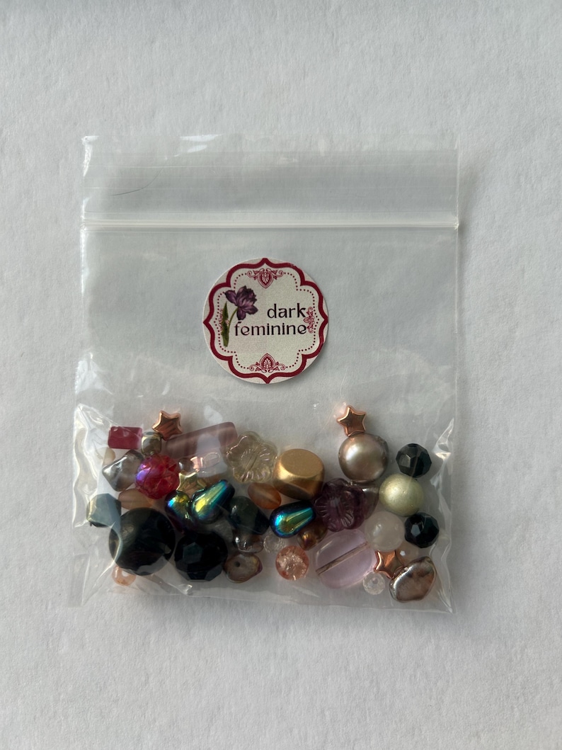 May include: A clear plastic resealable bag containing an assortment of loose beads in various shapes, sizes, and colors, including black, pink, gold, and iridescent tones. A circular sticker on the bag features a floral graphic and the text dark feminine.
