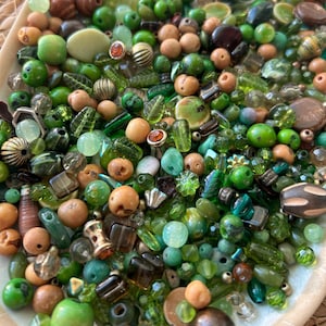 May include: A collection of assorted beads in various shades of green, brown, and tan. The mix includes round, leaf-shaped, faceted, and metallic beads made from glass, wood, and metal, featuring diverse textures, finishes, and sizes.