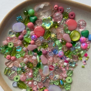 Puede incluir: Una colección de cuentas artesanales variadas en diversos tonos de rosa, verde y vidrio transparente. La mezcla incluye cuentas con forma de flor, cuentas con forma de hoja, esferas redondas y piezas ovaladas planas de diferentes tamaños.