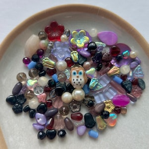 May include: A collection of assorted loose beads in various shapes, sizes, and colors, including translucent, opaque, and iridescent finishes. A central ceramic owl bead with brown, white, and blue details is surrounded by floral, round, and teardrop beads.