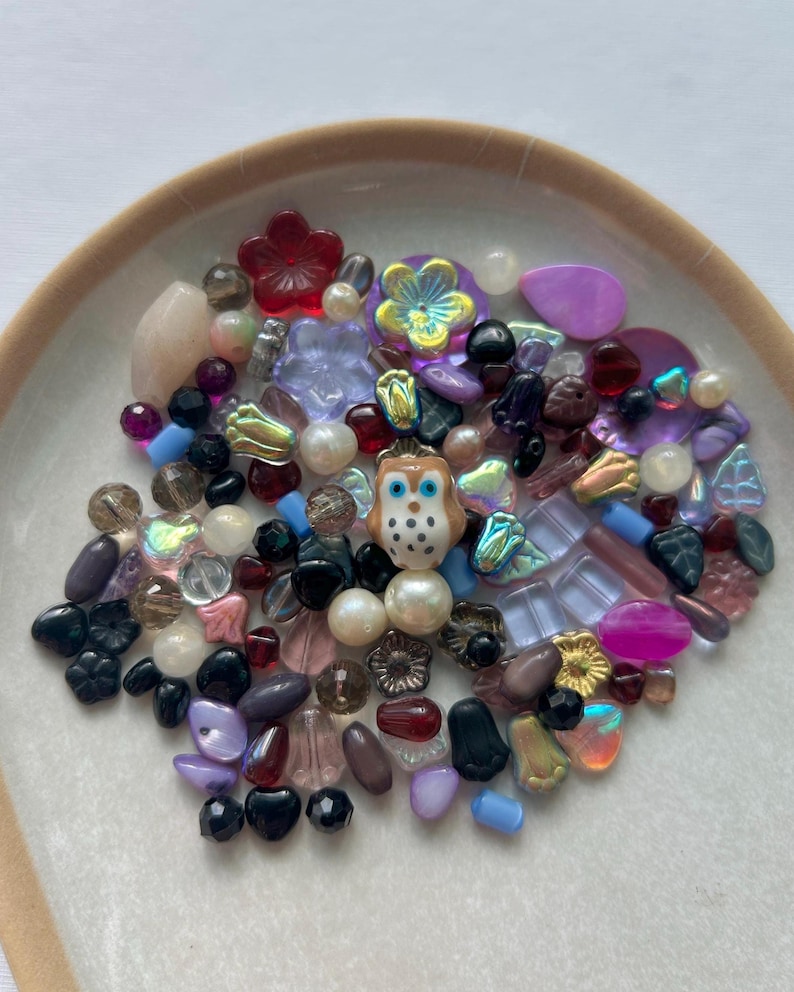 May include: A collection of assorted loose beads in various shapes, sizes, and colors, including translucent, opaque, and iridescent finishes. A central ceramic owl bead with brown, white, and blue details is surrounded by floral, round, and teardrop beads.
