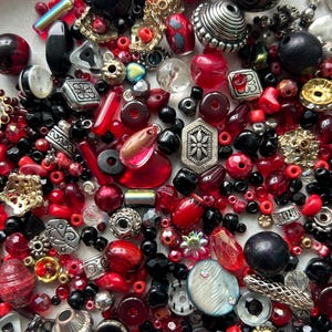 May include: A selection of beads in diverse forms, dimensions, and materials. Predominantly red, black, and silver, with touches of gold. The collection features round, faceted, and textured beads, ideal for jewellery making or crafting projects.