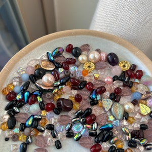 May include: A collection of assorted loose beads in various shapes, sizes, and colors including black, deep red, iridescent blue, clear, pink, and gold. The beads feature leaf, round, and teardrop forms with smooth, faceted, and metallic finishes.
