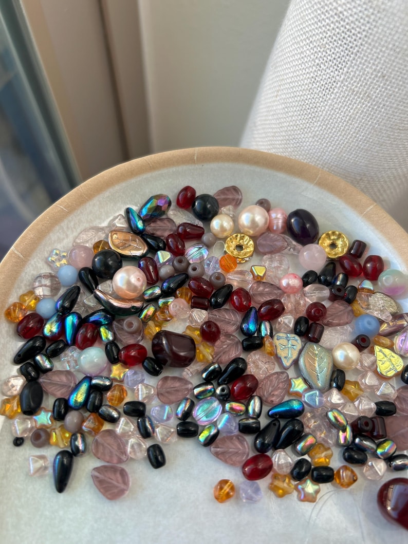 May include: A collection of assorted loose beads in various shapes, sizes, and colors including black, deep red, iridescent blue, clear, pink, and gold. The beads feature leaf, round, and teardrop forms with smooth, faceted, and metallic finishes.