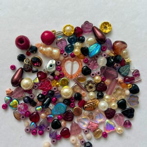 May include: A collection of assorted craft beads in various shapes, sizes, and textures. The mix includes round pearls, faceted crystals, leaf-shaped charms, heart-shaped beads, and metallic accents in shades of pink, purple, gold, and black.
