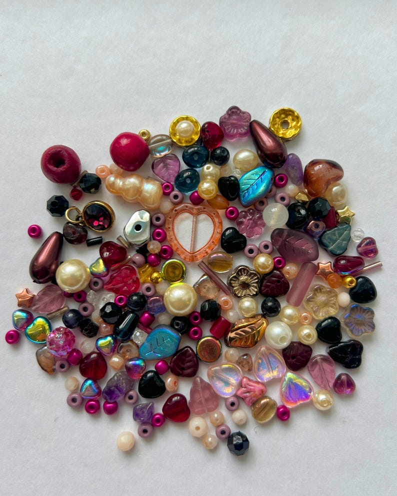 May include: A collection of assorted craft beads in various shapes, sizes, and textures. The mix includes round pearls, faceted crystals, leaf-shaped charms, heart-shaped beads, and metallic accents in shades of pink, purple, gold, and black.
