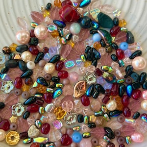 May include: A collection of assorted loose beads in various shapes, sizes, and colors, including round pearls, faceted crystals, leaf-shaped charms, and flower-shaped beads in shades of red, pink, blue, gold, and iridescent finishes.