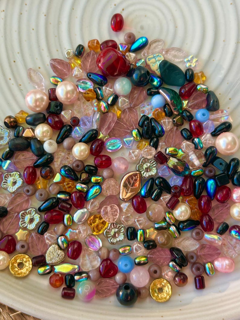 May include: A collection of assorted loose beads in various shapes, sizes, and colors, including round pearls, faceted crystals, leaf-shaped charms, and flower-shaped beads in shades of red, pink, blue, gold, and iridescent finishes.