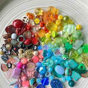 May include: A collection of colorful beads arranged on a white plate, showcasing a rainbow of hues. The beads vary in shape and size, including round, floral, and geometric designs. Colors range from red and orange to yellow, green, blue, and purple.