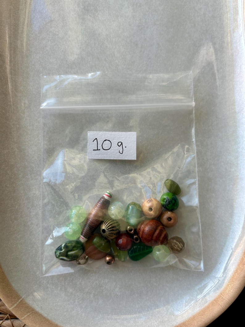 May include: A clear plastic resealable bag containing a collection of assorted beads in various shades of green, brown, and metallic tones. A white label inside the bag displays the handwritten text 10 g. The beads vary in shape, size, and texture.