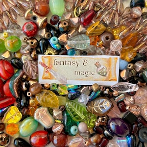 Può includere: Una vasta collezione di perline in vetro e pietra assortite in varie forme, dimensioni e colori, tra cui rosso, verde, ambra e nero. Un'etichetta rettangolare bianca al centro riporta il testo fantasy & magic con una spada e una pozione.