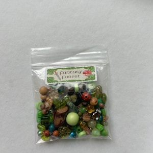 May include: A clear plastic resealable bag containing a variety of loose beads in shades of green, brown, and clear. The bag features a white label with green leaf borders, a small sword icon, a mushroom icon, and the text Fantasy Forest.
