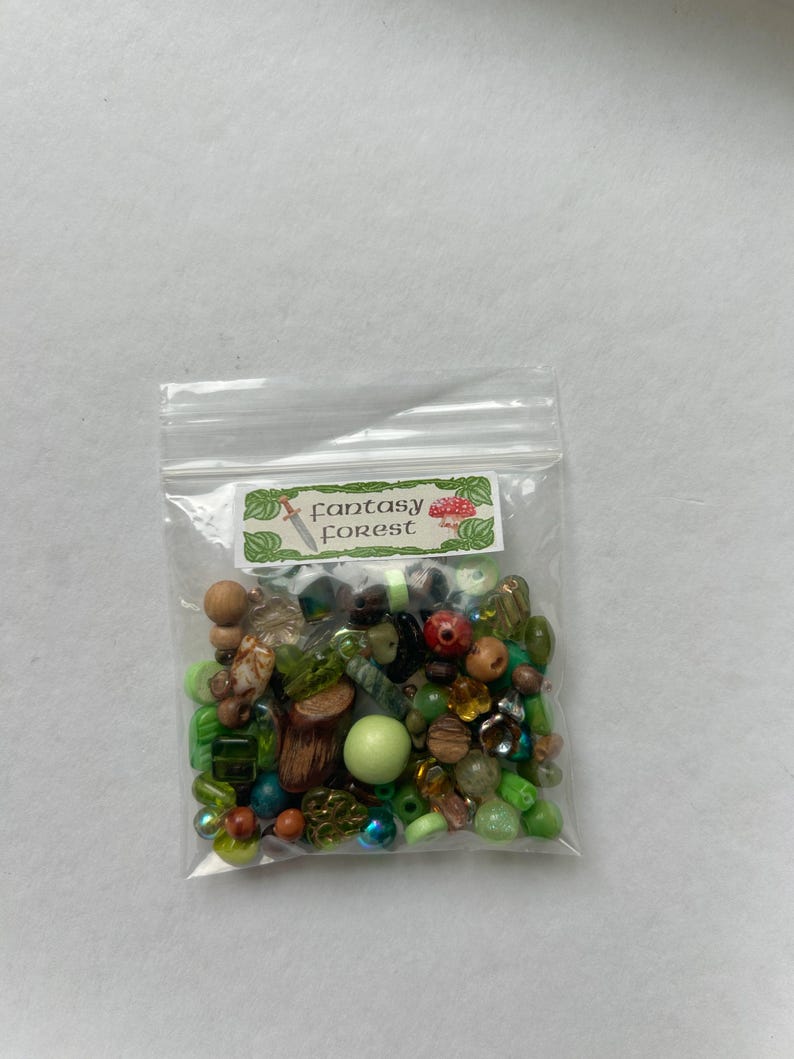 May include: A clear plastic resealable bag containing a variety of loose beads in shades of green, brown, and clear. The bag features a white label with green leaf borders, a small sword icon, a mushroom icon, and the text Fantasy Forest.