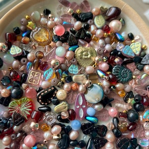 May include: A collection of assorted beads in a shallow bowl, featuring various shapes including leaves, hearts, and spheres. The beads are rendered in shades of pink, black, gold, blue, and clear glass, with iridescent, matte, and metallic finishes.