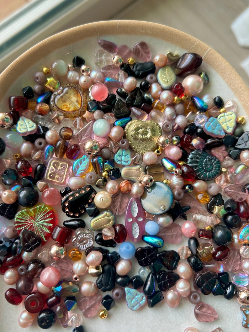 May include: A collection of assorted beads in a shallow bowl, featuring various shapes including leaves, hearts, and spheres. The beads are rendered in shades of pink, black, gold, blue, and clear glass, with iridescent, matte, and metallic finishes.