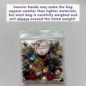 May include: A clear plastic bag filled with an assortment of multi-colored beads in various shapes and sizes. A circular white label with a purple flower graphic and the text "dark feminine" is attached. Text above reads: heavier beads may make the bag appear smaller than lighter materials, but each bag is carefully weighed and will always exceed the listed weight!