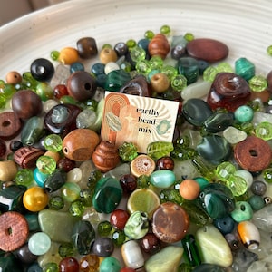 Puede incluir: Una mezcla de cuentas de diversas formas y colores, incluyendo verde, marrón y transparente. Las cuentas están dispersas sobre un plato blanco, con una pequeña tarjeta que dice "earthy bead mix".