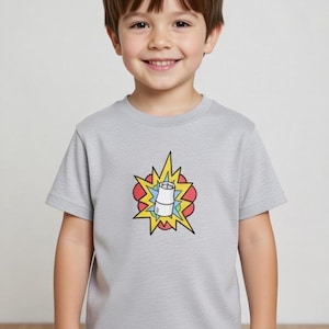 Puede incluir: Una camiseta gris claro de manga corta con un gráfico colorido de una explosión de dibujos animados y un objeto blanco en el centro. La camiseta la lleva un niño sonriente.