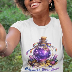 Puede incluir: Camiseta blanca con un diseño de botella de poción púrpura. La botella tiene un corcho, una cinta arcoíris y dijes de corazón. La botella tiene un diseño de galaxia. El texto "Polymorph Your Heart" está debajo.