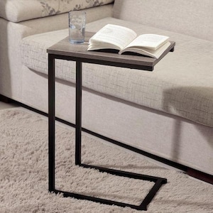 Pode incluir: Uma mesa lateral com um tampo cinza escuro e uma estrutura de metal preta. Um livro aberto e um copo transparente estão sobre a mesa. A mesa está ao lado de um sofá cinza claro e assenta sobre um tapete fofo e claro.