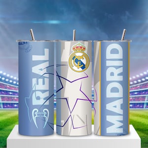 Puede incluir: Tres vasos térmicos del Real Madrid con pajitas plateadas. Los vasos presentan el logotipo del equipo, las palabras "REAL" y "MADRID" en blanco y un diseño de estrella. El fondo muestra un estadio con luces brillantes y un campo verde.