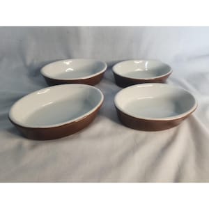 May include: A set of four oval-shaped baking dishes featuring a smooth white interior and a solid brown exterior. Each dish has a shallow depth and a rounded rim, designed with a consistent colour scheme and uniform shape for kitchen use.